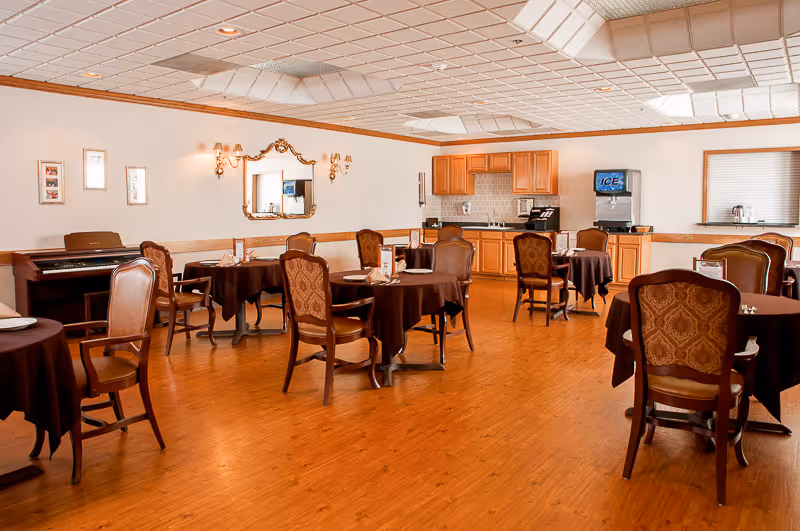 A dining room with several round tables covered with dark tablecloths, each surrounded by wooden chairs with patterned upholstery. The room has wooden flooring, a piano against one wall, a mirror with wall sconces, and a kitchenette area with cabinets, a coffee maker, and an ice dispenser.
