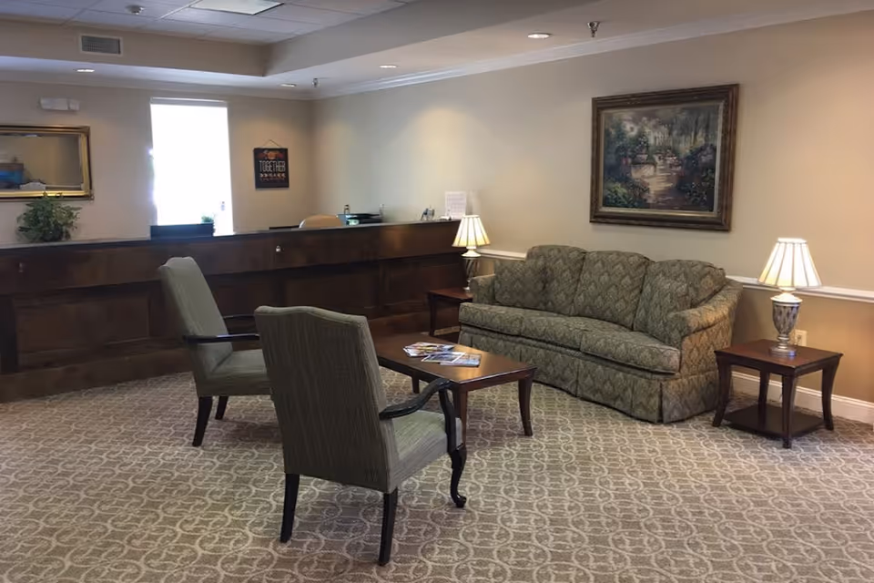 A cozy sitting area in a senior living facility with a patterned sofa, two upholstered chairs, a wooden coffee table with magazines, two side tables with lamps, a framed painting on the wall, and a wooden reception desk in the background.