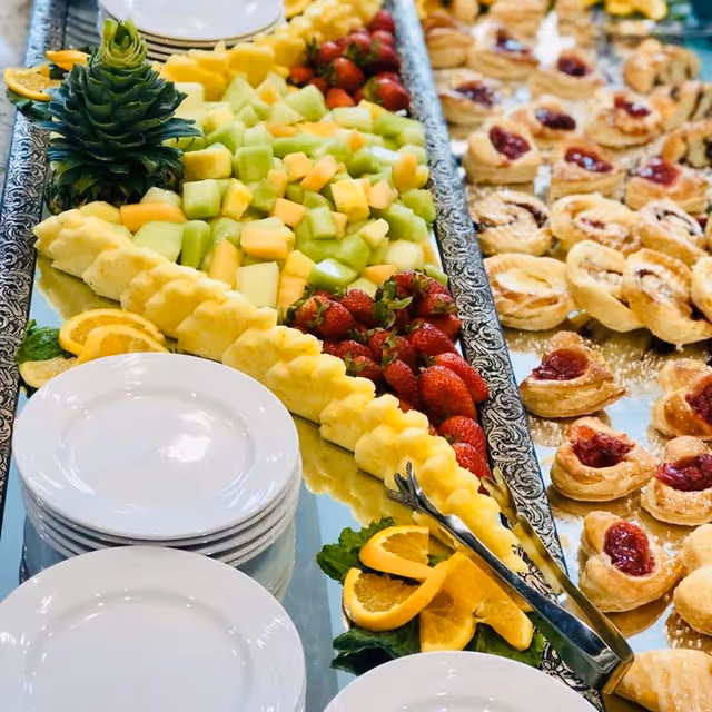 A buffet table with a decorative tray of fresh fruit including pineapple, strawberries, cantaloupe, and honeydew melon, alongside a stack of white plates and a tray of small pastries with fruit filling.