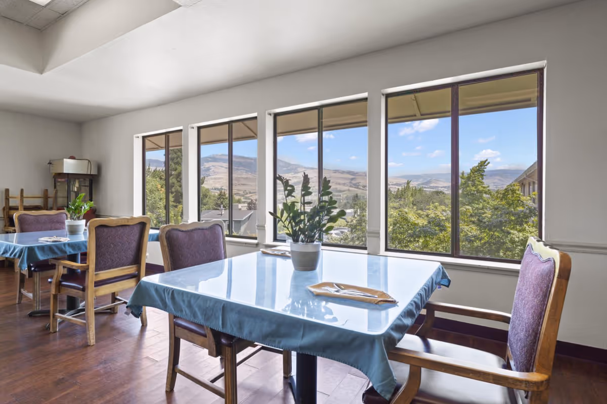 Sunlit dining room with tables and upholstered chairs by large windows showing a hilly landscape.