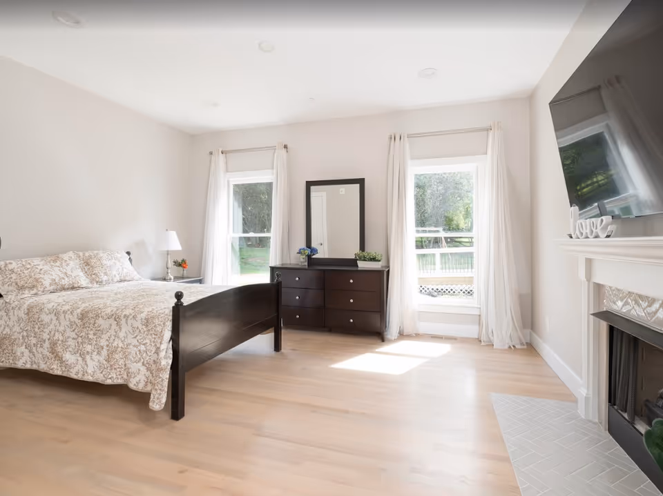 A bright and airy bedroom with light wood flooring, a dark wooden bed with floral bedding, a matching dresser with a mirror between two windows with sheer white curtains, a bedside table with a lamp and flowers, and a fireplace with a mounted TV above it and a decorative 'love' sign on the mantel.