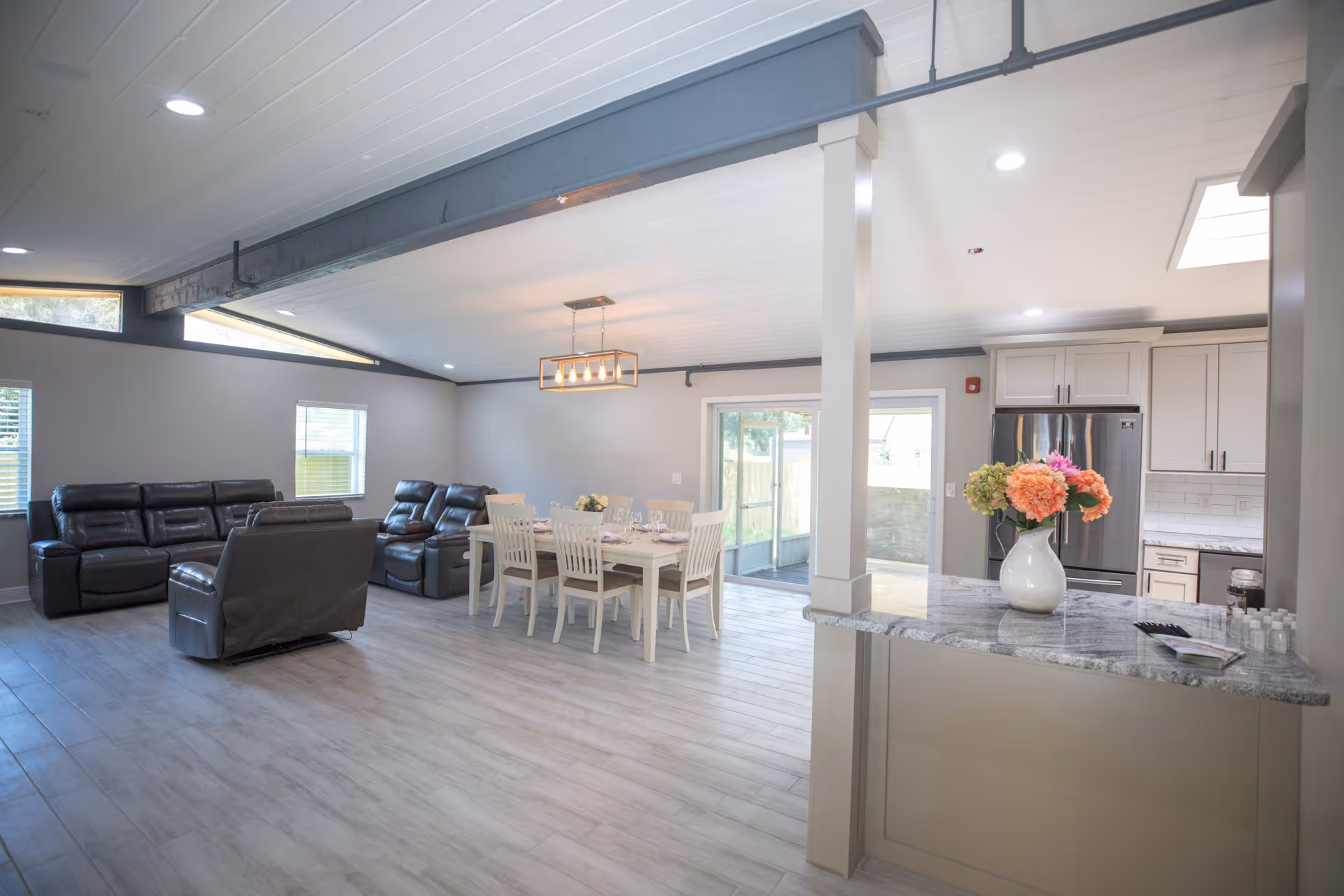A spacious open-concept living and dining area in an assisted living facility. The room features light gray walls and wood-look flooring. There are three dark leather recliners arranged in the living area, and a white dining table with six chairs set for a meal. The kitchen area is visible with light-colored cabinets, a stainless steel refrigerator, and a granite countertop with a vase of colorful flowers. Large windows and a sliding glass door allow natural light to enter the space.