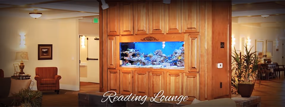 Interior view of a reading lounge featuring a large built-in aquarium surrounded by wooden cabinetry. The room includes comfortable seating such as armchairs and sofas, warm lighting fixtures, and decorative plants, creating a cozy and inviting atmosphere.