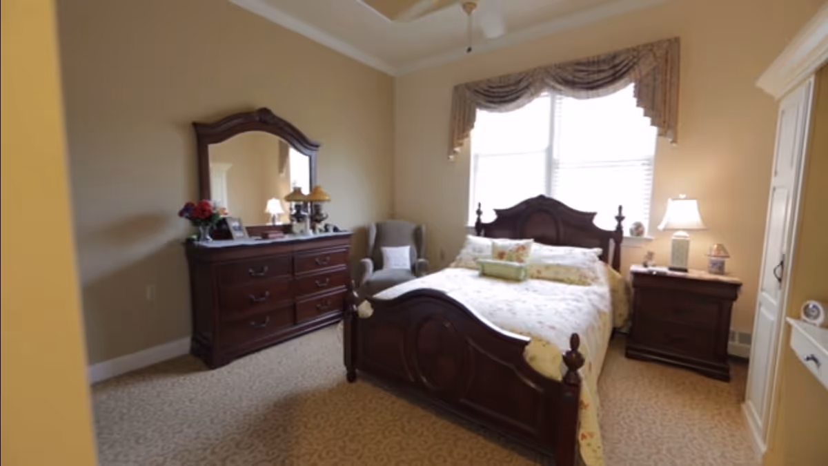 A cozy bedroom with a wooden bed frame, floral bedding, a matching wooden dresser with a large mirror, a nightstand with a lamp, and a comfortable armchair near a window with decorative curtains.