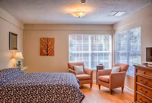 A cozy bedroom with a large bed covered in a dark blue and white patterned bedspread. There is a nightstand with a lamp and a small picture frame next to the bed. Two cushioned armchairs with light-colored pillows are positioned near large windows with white blinds, allowing natural light to fill the room. A wooden dresser with a TV on top is on the right side of the room. A framed artwork depicting a tree hangs on the wall above the armchairs.