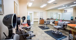 Interior view of a salon area in a senior living facility with multiple salon chairs, hair dryers, mirrors, and a water dispenser. The room has beige walls, tiled floors, and bright overhead lighting.