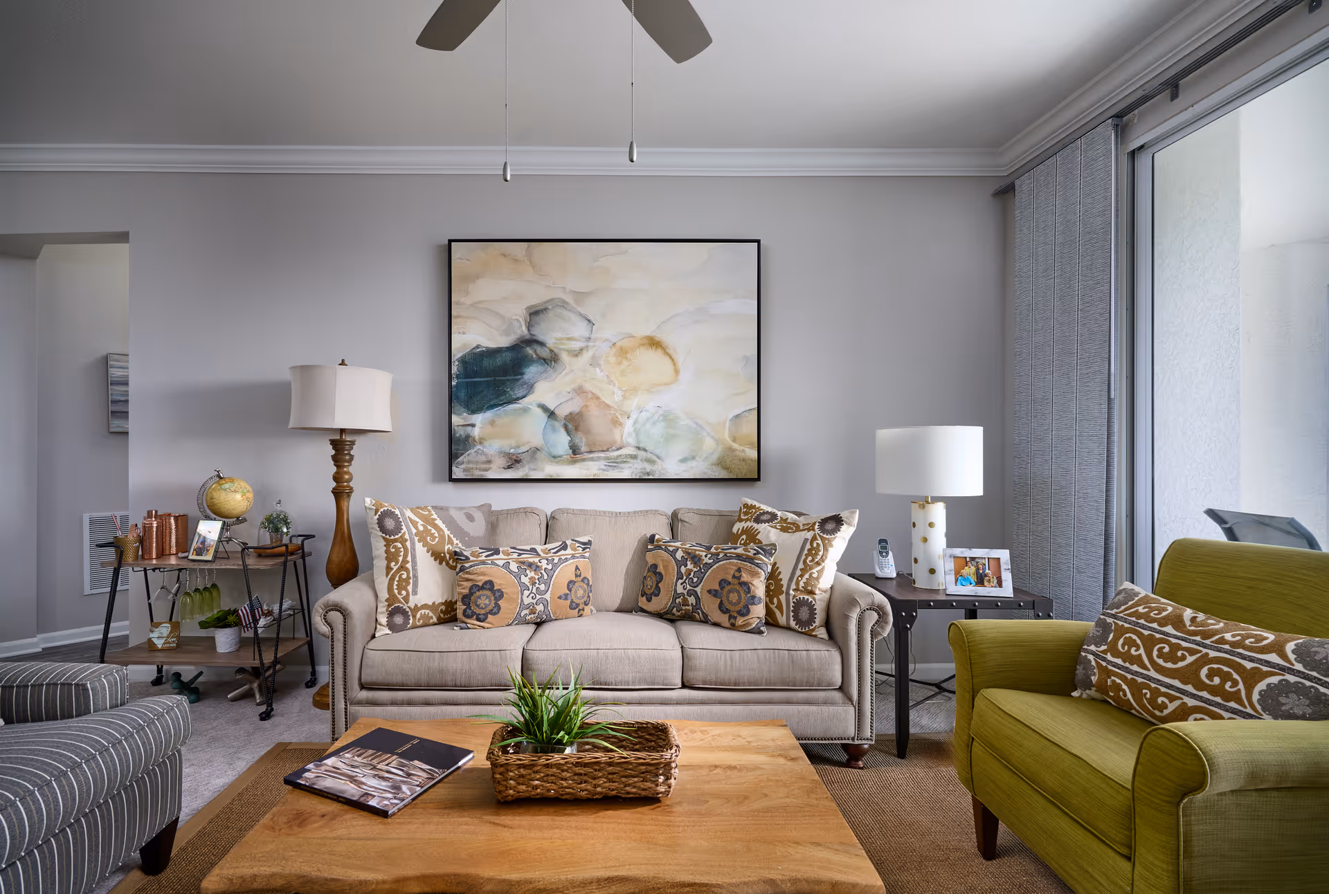 Well-lit living room with a beige sofa, patterned throw pillows, a green armchair, coffee table and large abstract painting on the wall.