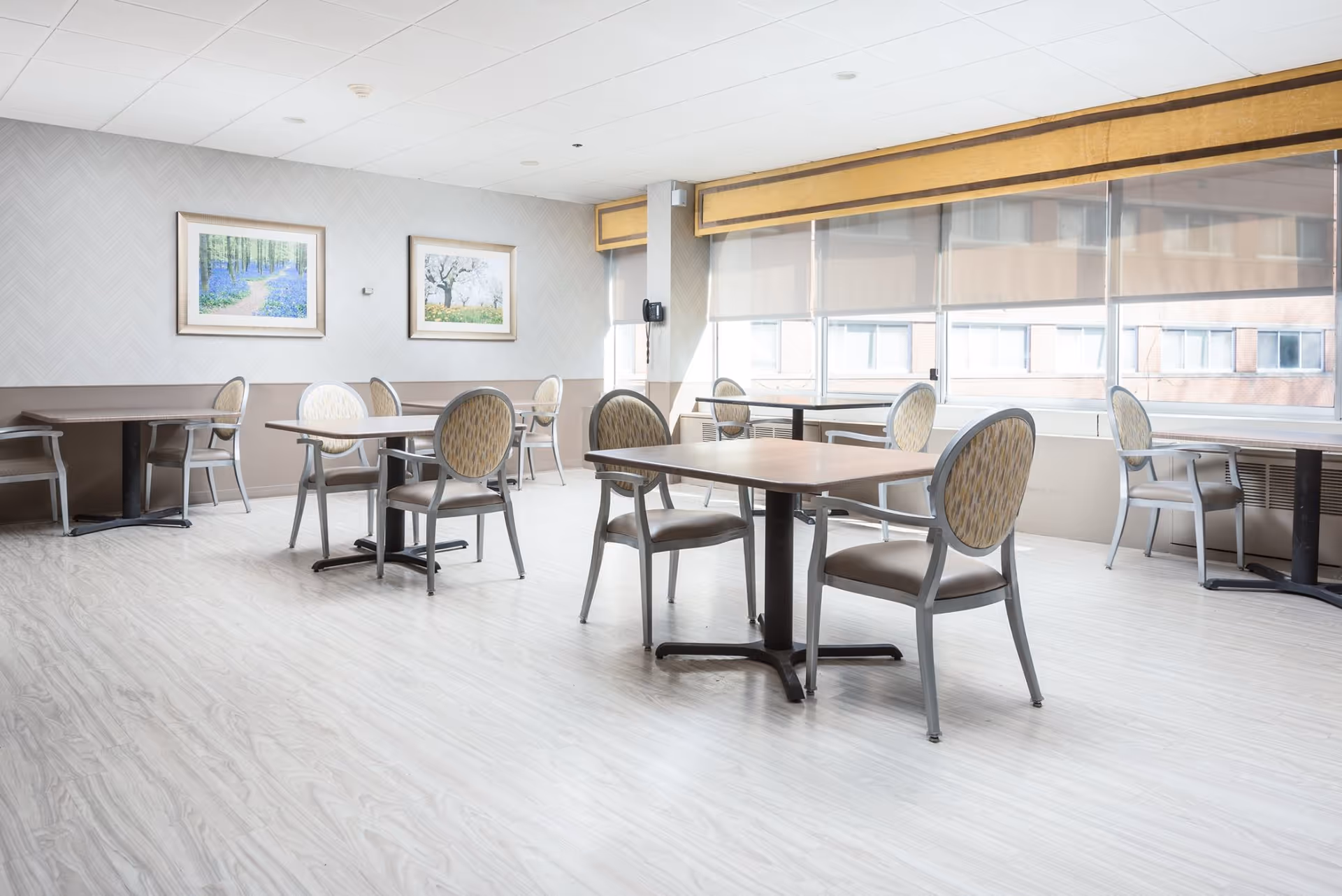 Bright communal dining room with tables and chairs, large windows, and framed artwork on the walls.