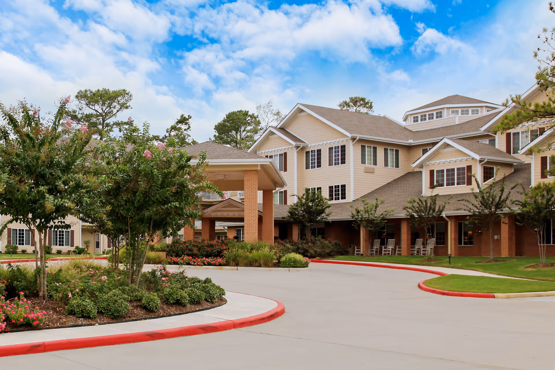 Exterior view of a senior living facility with beige siding and brick accents, surrounded by landscaped greenery and trees under a partly cloudy blue sky. The driveway curves around a garden bed with flowering plants leading to a covered entrance with columns.