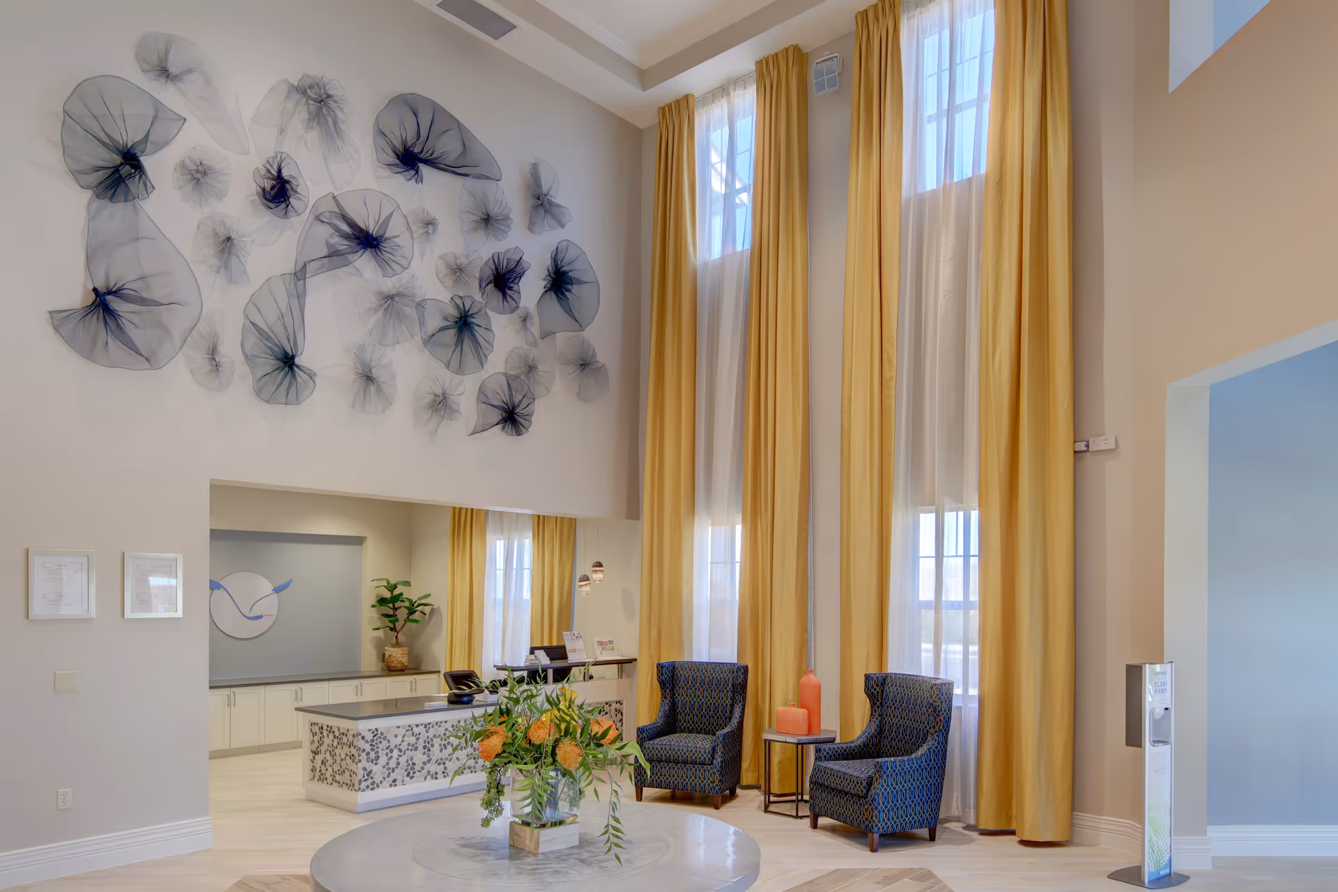 A bright and spacious assisted living facility lobby with tall windows covered by long yellow curtains. Two patterned armchairs are placed near a small round table with decorative items. A large round table with a floral arrangement is in the foreground. The wall features artistic black mesh decorations, and a reception desk is visible in the background.
