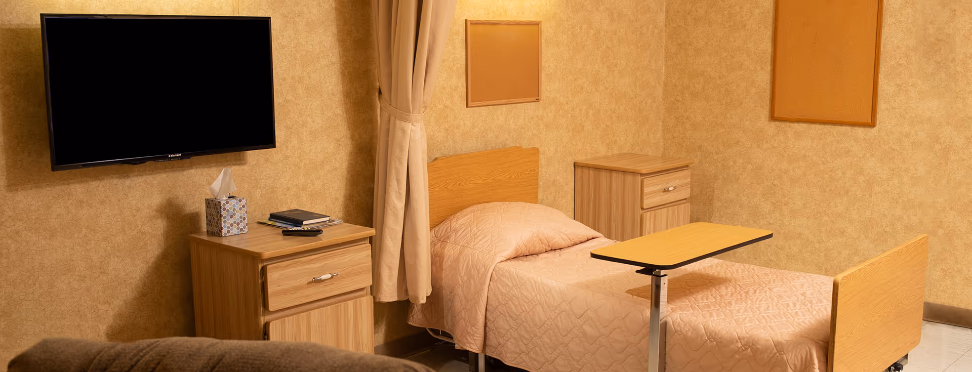 A simple bedroom in a senior living facility featuring a single bed with a light pink quilted bedspread, a wooden headboard and footboard, a small adjustable overbed table, two wooden nightstands with drawers, a mounted flat-screen TV on the wall, a box of tissues, and a remote control. The walls are beige with two empty cork bulletin boards and a beige curtain partially visible.