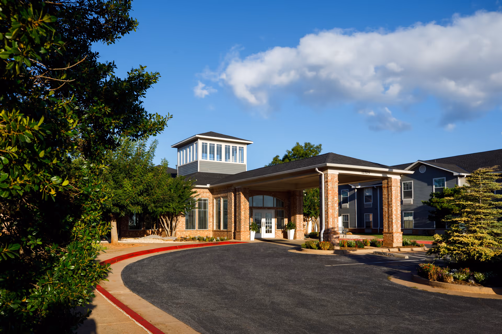 Exterior view of Acclaim Living facility showing the main entrance with a covered drop-off area supported by brick columns, surrounded by trees and landscaping under a blue sky with some clouds.