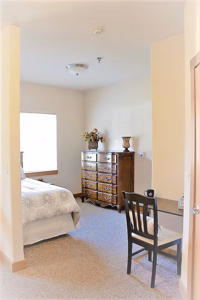 A bright bedroom with a bed covered in a white patterned bedspread, a wooden dresser with decorative items on top, and a small desk with a chair and a coffee mug. The room has beige walls and carpeted floor, with a window letting in natural light.