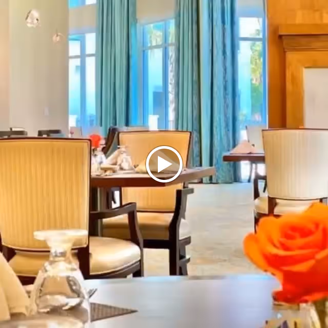 A bright dining room with large windows covered by blue curtains, several tables set with glassware and napkins, and beige upholstered chairs. An orange flower is visible in the foreground on one of the tables.