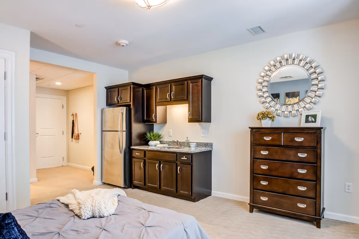 Interior view of a senior living facility room at Arbor Terrace Mountainside featuring a small kitchenette with dark wood cabinets, a stainless steel refrigerator, a granite countertop with a sink, and a potted plant. To the right, there is a dark wood chest of drawers with a decorative round mirror above it and a framed photo and flower pot on top. The room has light-colored walls and carpeted flooring, with a bed partially visible in the foreground.