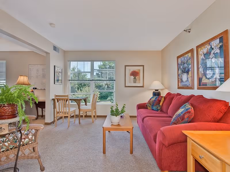 A cozy living room with a red sofa adorned with colorful pillows, a wooden coffee table with a potted plant, and framed floral artwork on the walls. There is a window letting in natural light, a small dining table with two chairs near the window, and a wicker chair with a green potted plant on a side table.
