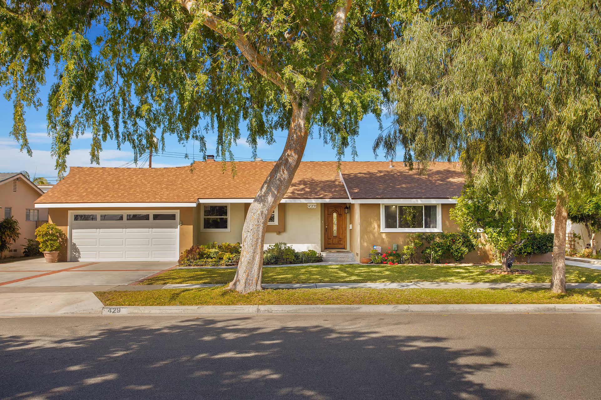 Single-story residential house with a brown shingle roof, beige exterior walls, a wooden front door, and a white garage door. The house is surrounded by a well-maintained lawn, shrubs, and trees, with a sidewalk and street in the foreground under a clear blue sky.