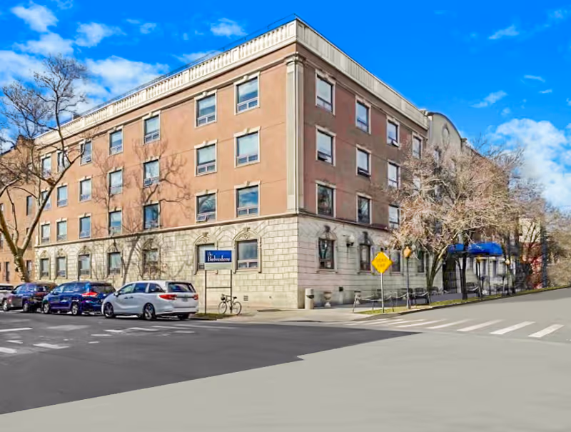 Four-story brick-and-stone senior living building on a tree-lined street corner with parked cars and a blue entrance awning.