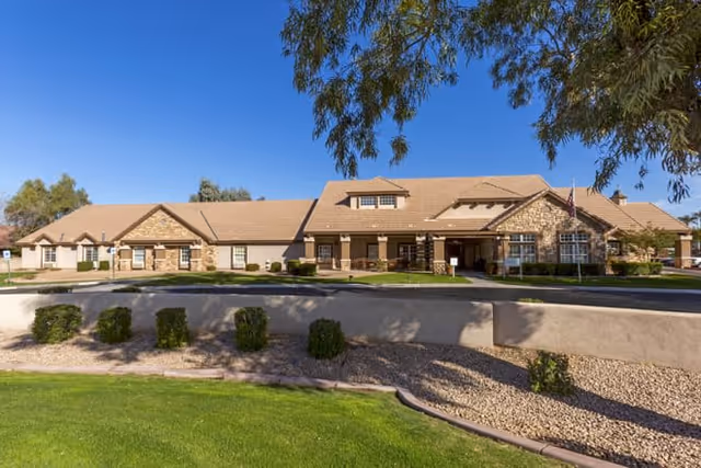 Exterior view of a single-story senior living facility building with a tan roof and stone accents, surrounded by green grass, trees, and landscaping under a clear blue sky.
