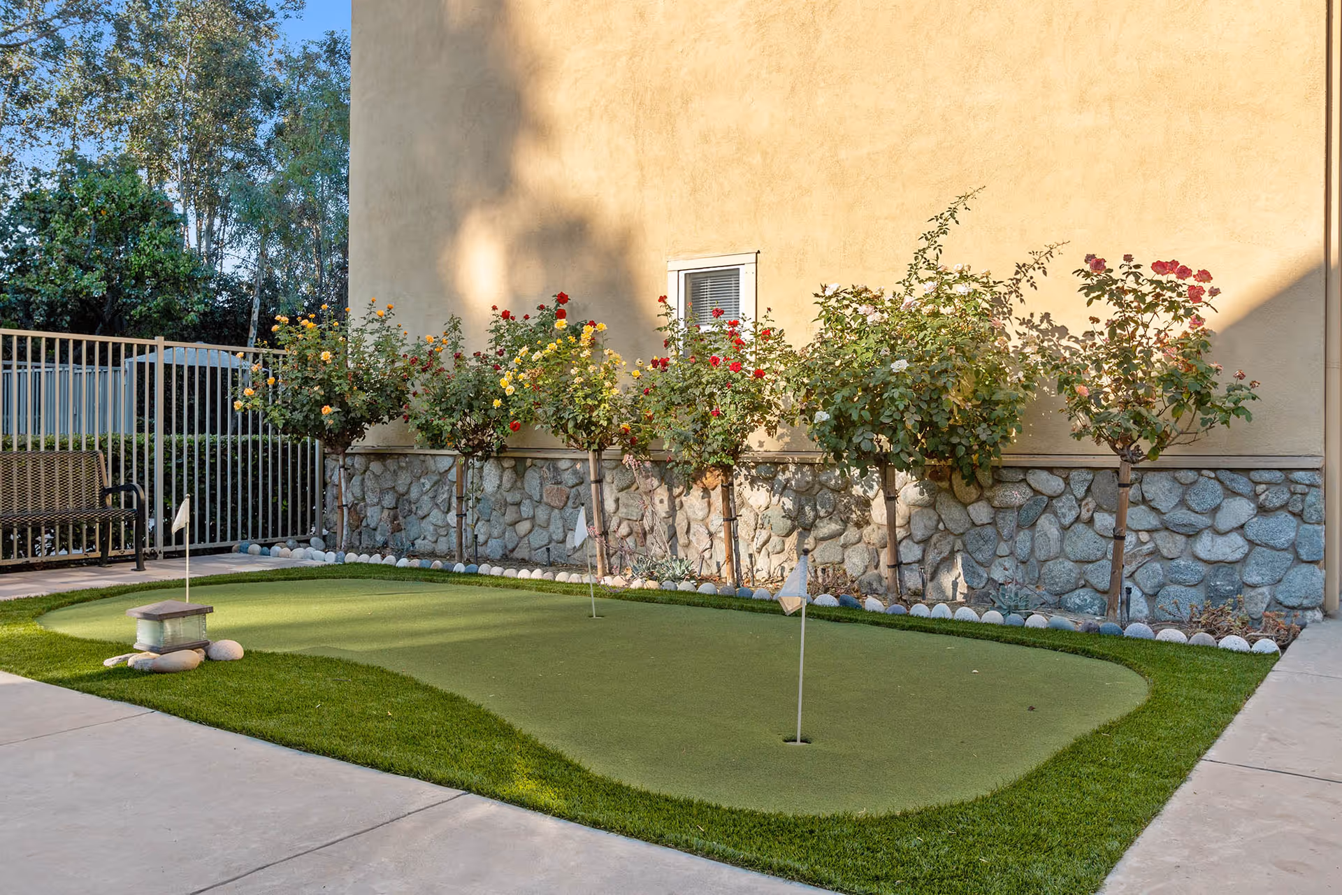 Small outdoor putting green with three holes and flags, surrounded by a concrete walkway. There is a row of rose bushes with red, yellow, and orange flowers along a stone and stucco wall. A metal bench is visible near a metal fence in the background.
