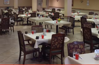 A spacious dining room with multiple tables covered in white tablecloths, each set with glasses filled with red beverages, salt and pepper shakers, and small flower arrangements. The room has tiled floors and upholstered chairs with patterned fabric.