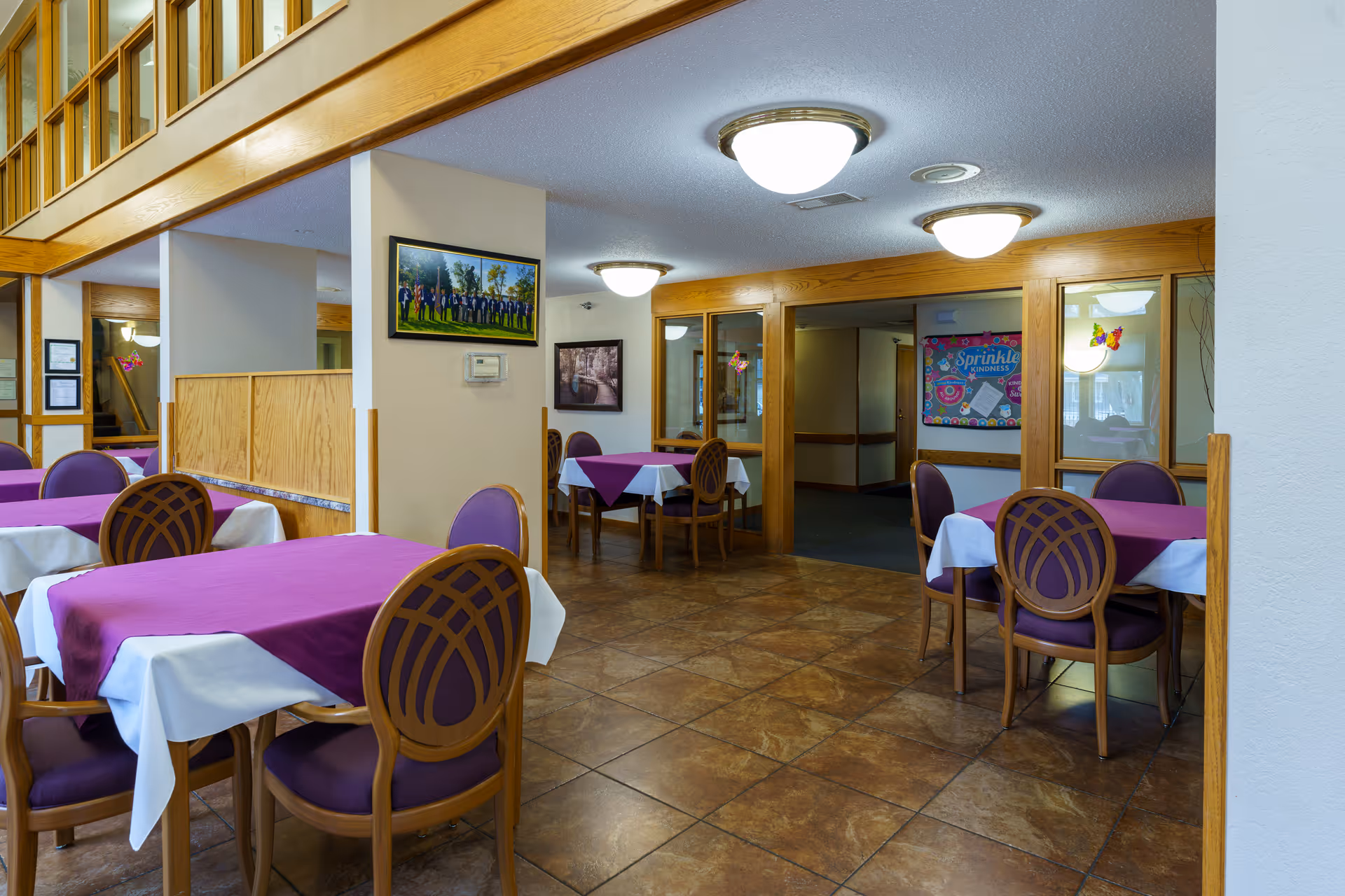 Interior view of a dining area with multiple tables covered in white tablecloths and purple table runners. Each table is surrounded by wooden chairs with purple cushions. The room has tiled flooring, wooden trim, and ceiling lights. There are framed pictures on the walls and a bulletin board in the background.