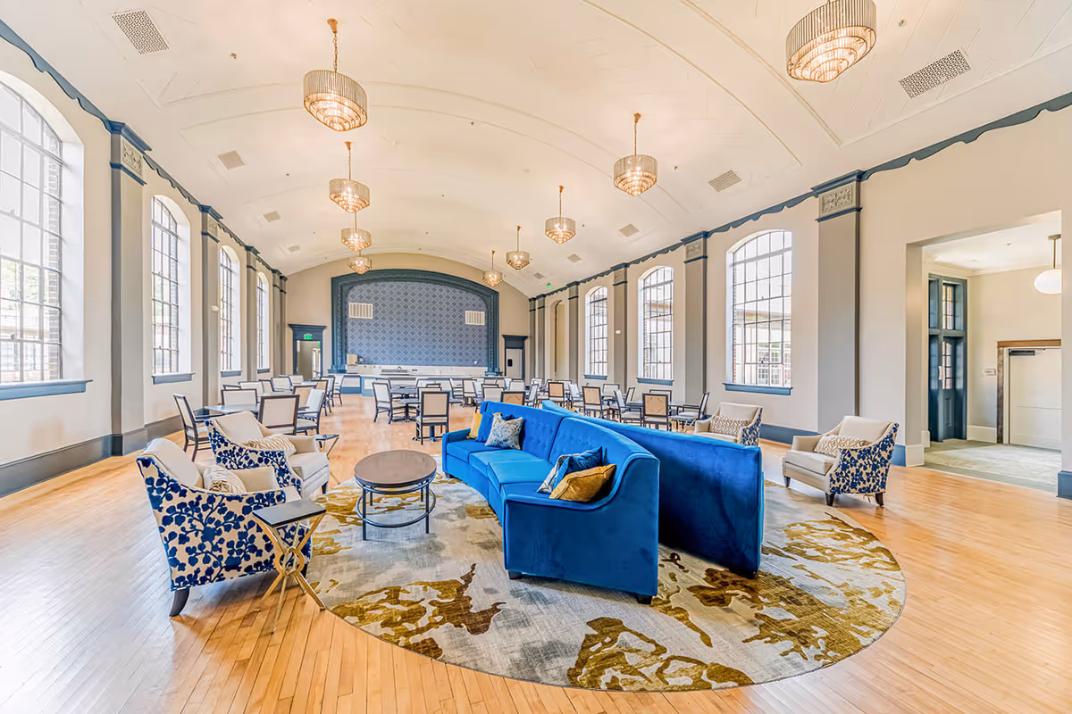 Spacious and elegant common area with high vaulted ceilings and large arched windows allowing natural light to fill the room. The room features a blue velvet sofa with decorative pillows, patterned armchairs, a round coffee table on a large area rug, and multiple dining tables with chairs arranged towards the back. Several chandeliers hang from the ceiling, and the walls are painted in neutral tones with blue trim accents.