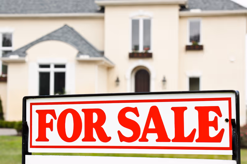 A 'For Sale' sign in bold red letters is placed in front of a large beige house with multiple windows and a dark front door.