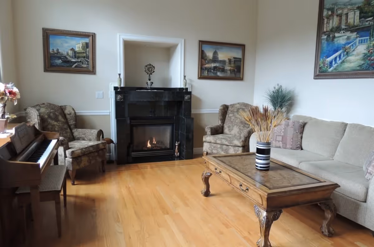 A cozy living room with a beige sofa, two patterned armchairs, a wooden coffee table with a striped vase holding dried plants, a black marble fireplace with a small fire burning, and a wooden piano with a bench. The walls are decorated with framed paintings depicting scenic views.