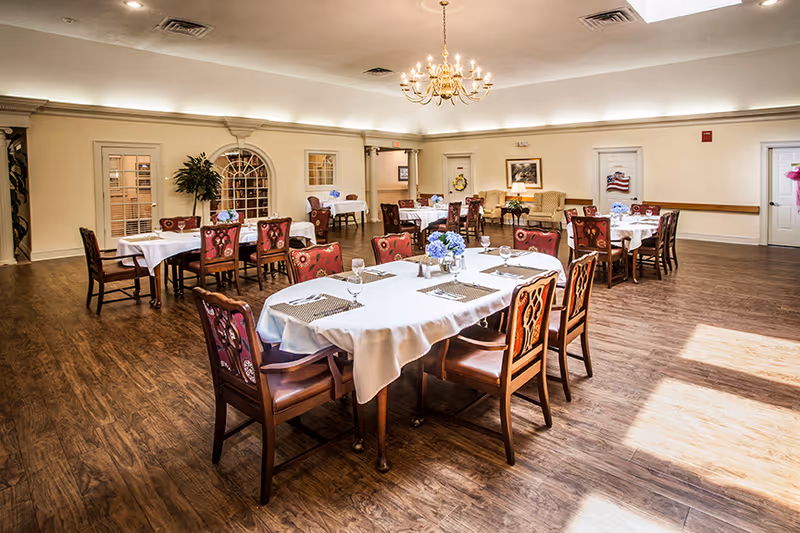 A spacious dining room with several tables covered in white tablecloths, each set with placemats, glasses, and floral centerpieces. The room has wooden flooring, cream-colored walls, and a chandelier hanging from the ceiling. There are upholstered chairs around the tables and some armchairs along the back wall near framed artwork and a door with an American flag decoration.