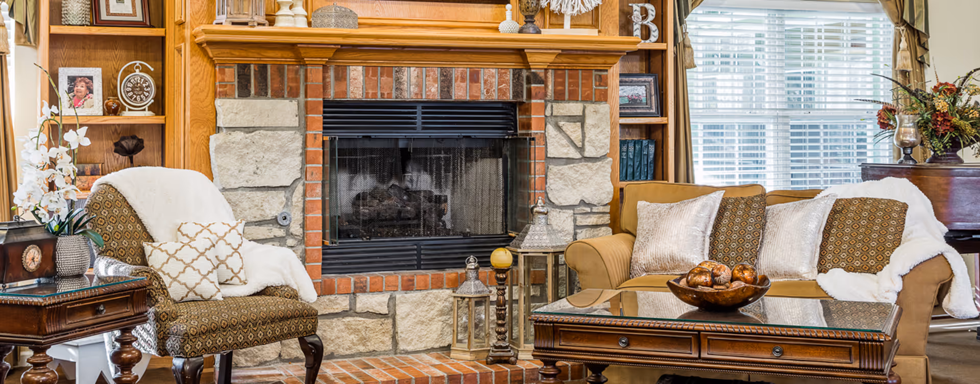 Cozy living room with a stone and brick fireplace in the center, flanked by wooden built-in shelves with decorative items and framed photos. A patterned armchair with white throw and pillows is on the left, and a beige sofa with multiple pillows and a white throw is on the right. A wooden coffee table with a decorative bowl sits in front of the sofa, and a large window with blinds and curtains is in the background.