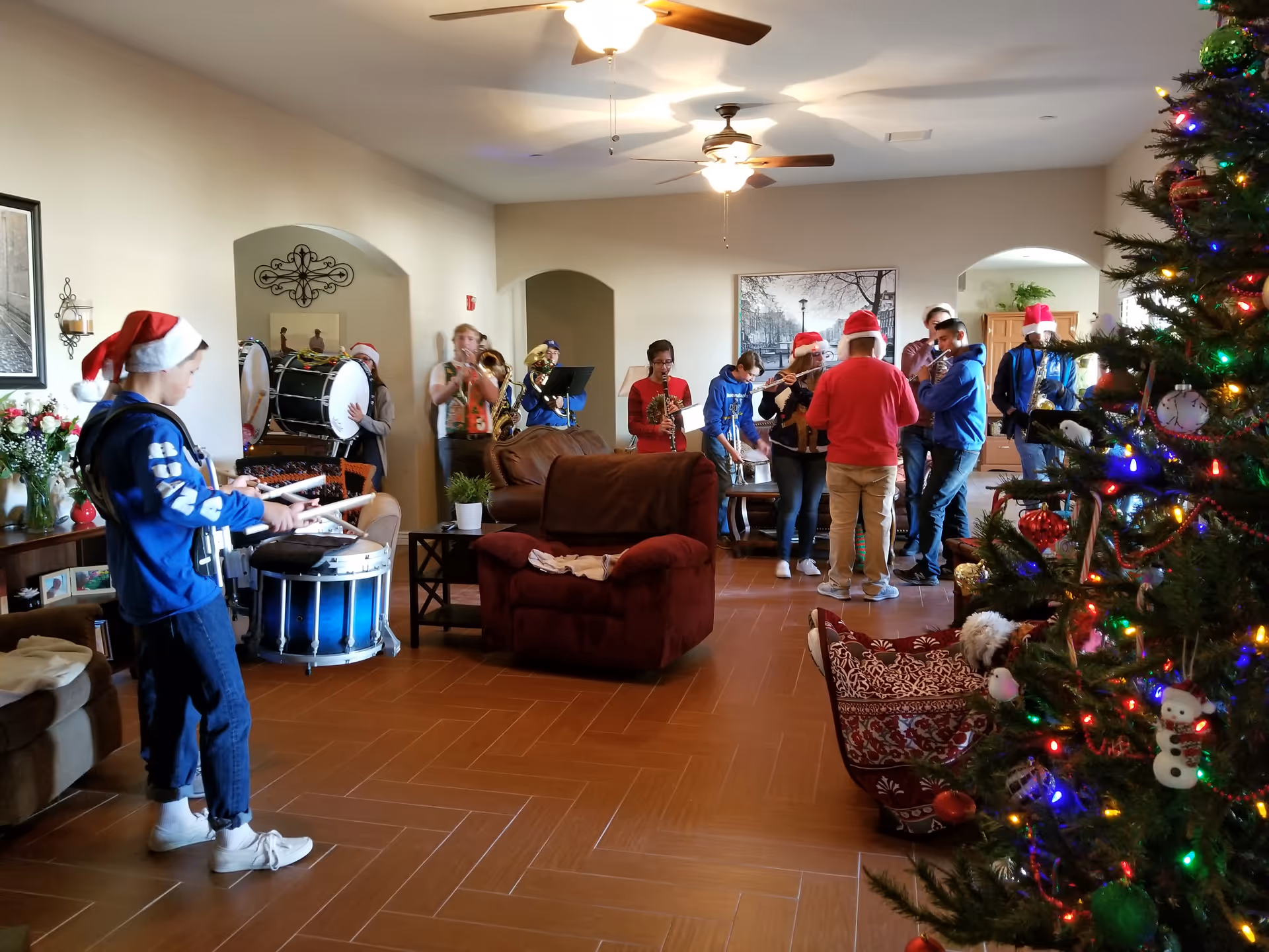 A group of people wearing Santa hats playing musical instruments in a living room decorated for Christmas with a Christmas tree adorned with colorful lights and ornaments. The room has ceiling fans, brown tile flooring, and comfortable seating including a brown armchair and sofa.