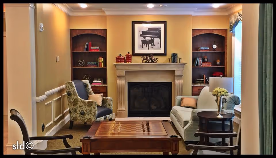 A cozy living room with a fireplace in the center, flanked by two built-in wooden bookshelves filled with books and decorative items. In front of the fireplace is a wooden coffee table with a chessboard on top. There are two armchairs with floral upholstery on the left and a light-colored sofa with pillows on the right. A small round side table with a vase of flowers is next to the sofa. The walls are painted a warm beige color, and there is a framed black and white picture of a piano above the fireplace.
