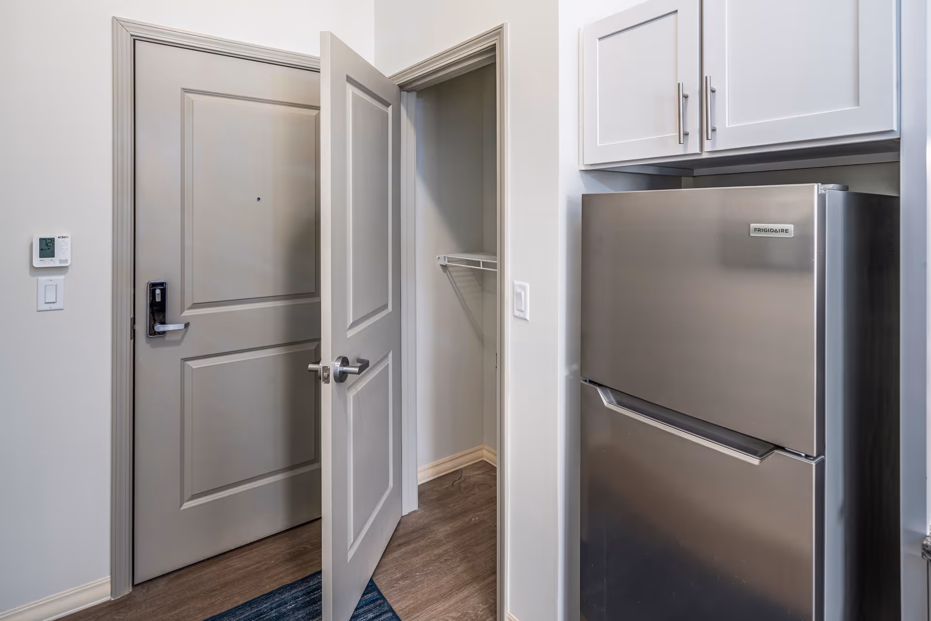 Interior view of a small kitchen area showing a stainless steel Frigidaire refrigerator, white cabinets above it, an open door revealing a small closet with a wire shelf, and a closed main entrance door with a digital thermostat on the wall beside it.