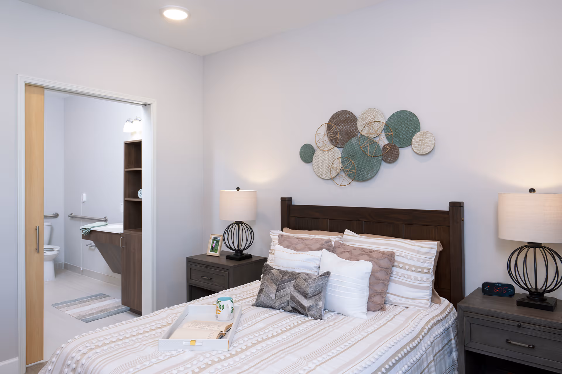 A cozy bedroom in Sage Valley Senior Living featuring a neatly made bed with multiple pillows and a tray holding an open book and a mug. On either side of the bed are matching nightstands with modern lamps. A decorative wall art piece with circular patterns hangs above the headboard. An open door reveals an adjacent bathroom with a toilet, sink, and grab bars.