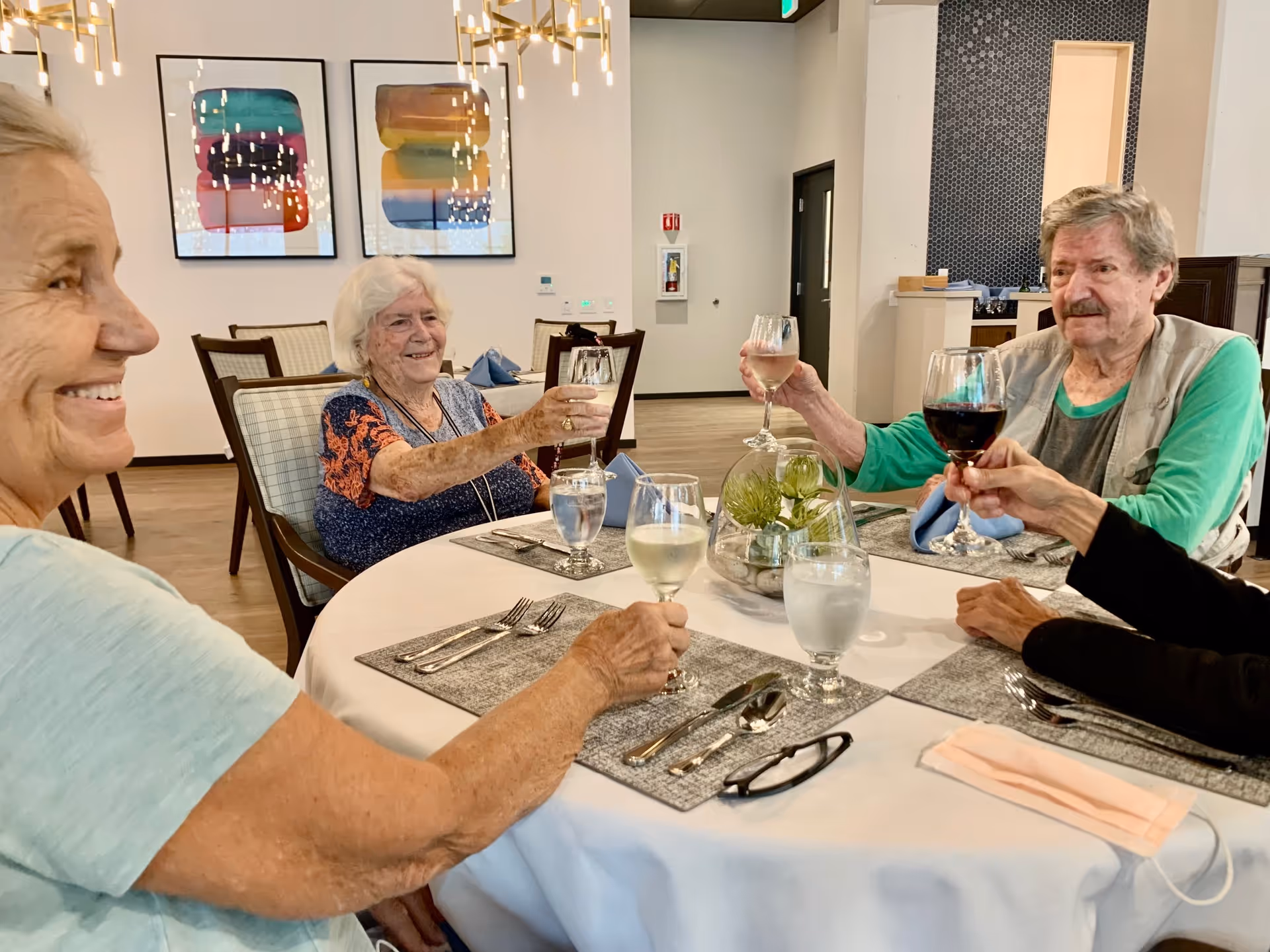Four elderly people sitting around a round dining table in a well-lit room, raising their wine glasses in a toast. The table is set with placemats, silverware, glasses of water, and a small decorative centerpiece. Colorful abstract paintings hang on the wall in the background.