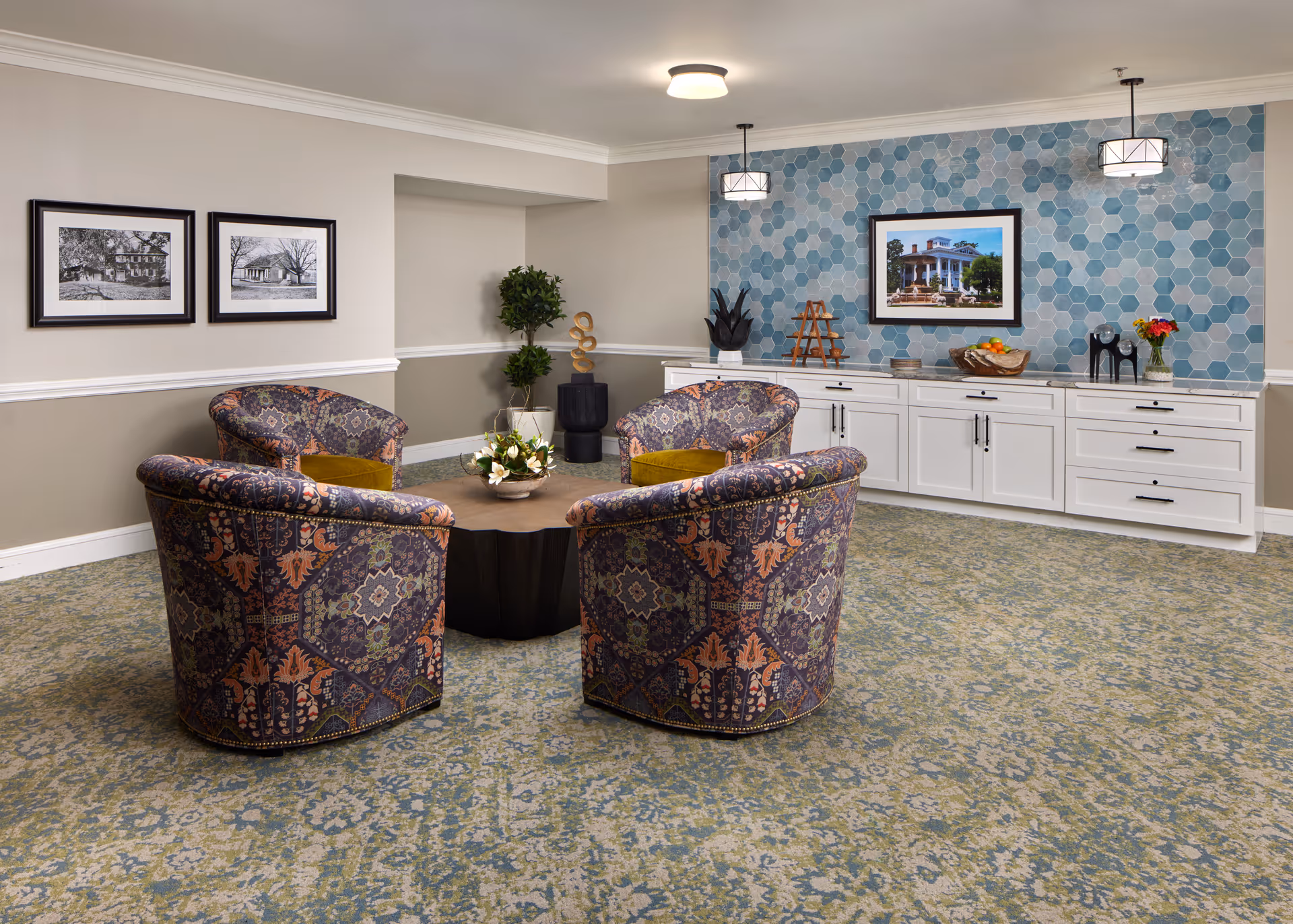 A cozy sitting area in a senior living facility with four patterned armchairs arranged around a round wooden coffee table. The room features a blue hexagonal tile accent wall with a framed picture of a building, white cabinetry with decorative items, two pendant lights, and framed black-and-white photos on a beige wall. The floor is covered with a patterned carpet.