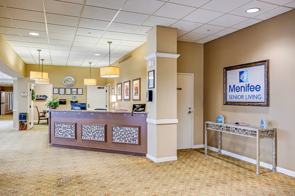 Reception lobby of Menifee Senior Living showing a front desk, hanging lights, seating, and a framed facility sign.