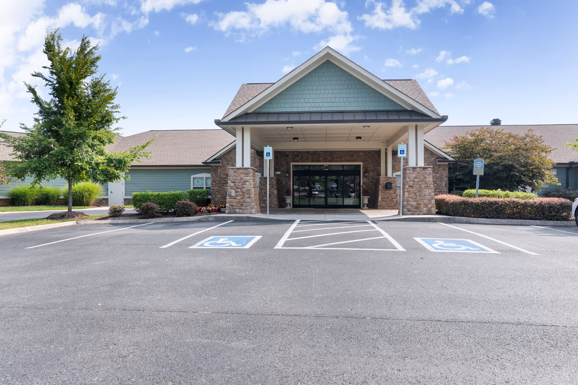 Covered main entrance of a single-story senior living facility with stone columns, glass doors, and marked handicap parking spaces in front.