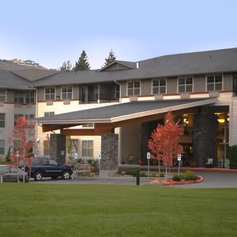 Exterior view of a senior living facility building with a covered entrance supported by stone pillars. There are a few small trees with red leaves and parked cars near the entrance. The building has multiple windows and a gray roof, with a grassy area in the foreground.