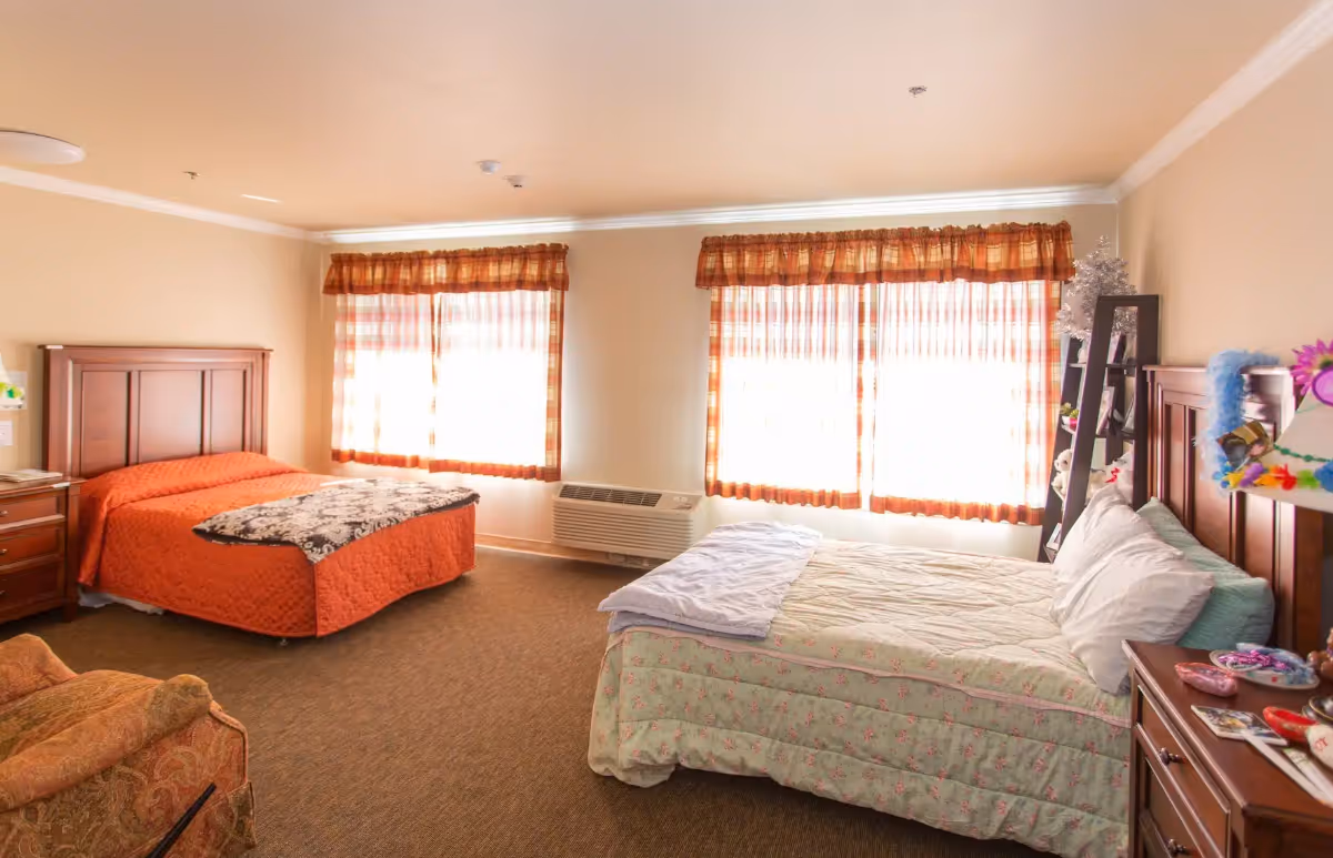 A well-lit bedroom with two beds, one with an orange bedspread and the other with a light floral bedspread. The room has two large windows with orange plaid curtains, a wooden nightstand, a wooden dresser, and a comfortable armchair. The walls are painted beige and the carpet is brown.