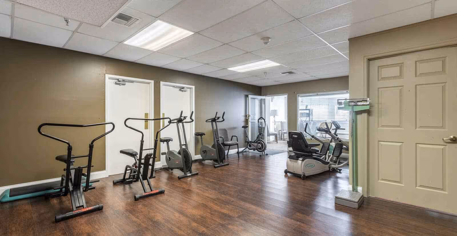 A fitness room with several exercise bikes and a recumbent bike on a wooden floor. There is a green scale near a beige door on the right, and two white doors on the left wall. The room has a drop ceiling with fluorescent lights and a window looking into another room with chairs and a lamp.