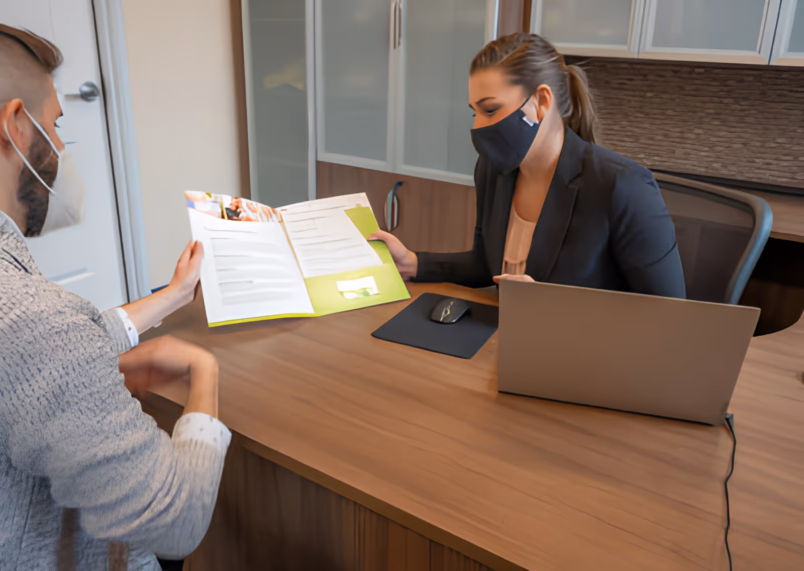 A woman wearing a black face mask and black blazer sits behind a wooden desk with a laptop and mouse, showing a brochure to a man wearing a white face mask and a light gray sweater. They appear to be in an office setting with cabinets in the background.