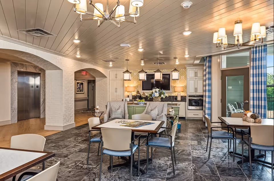 A well-lit, spacious common area in a senior living facility featuring multiple tables and chairs arranged for social or recreational activities. The room has a tiled floor, a ceiling with recessed lighting, and a kitchen area with cabinets, a countertop, and a mounted TV. There are two armchairs with pillows near the kitchen, blue and white checkered curtains by a glass door leading outside, and decorative lighting fixtures on the ceiling.