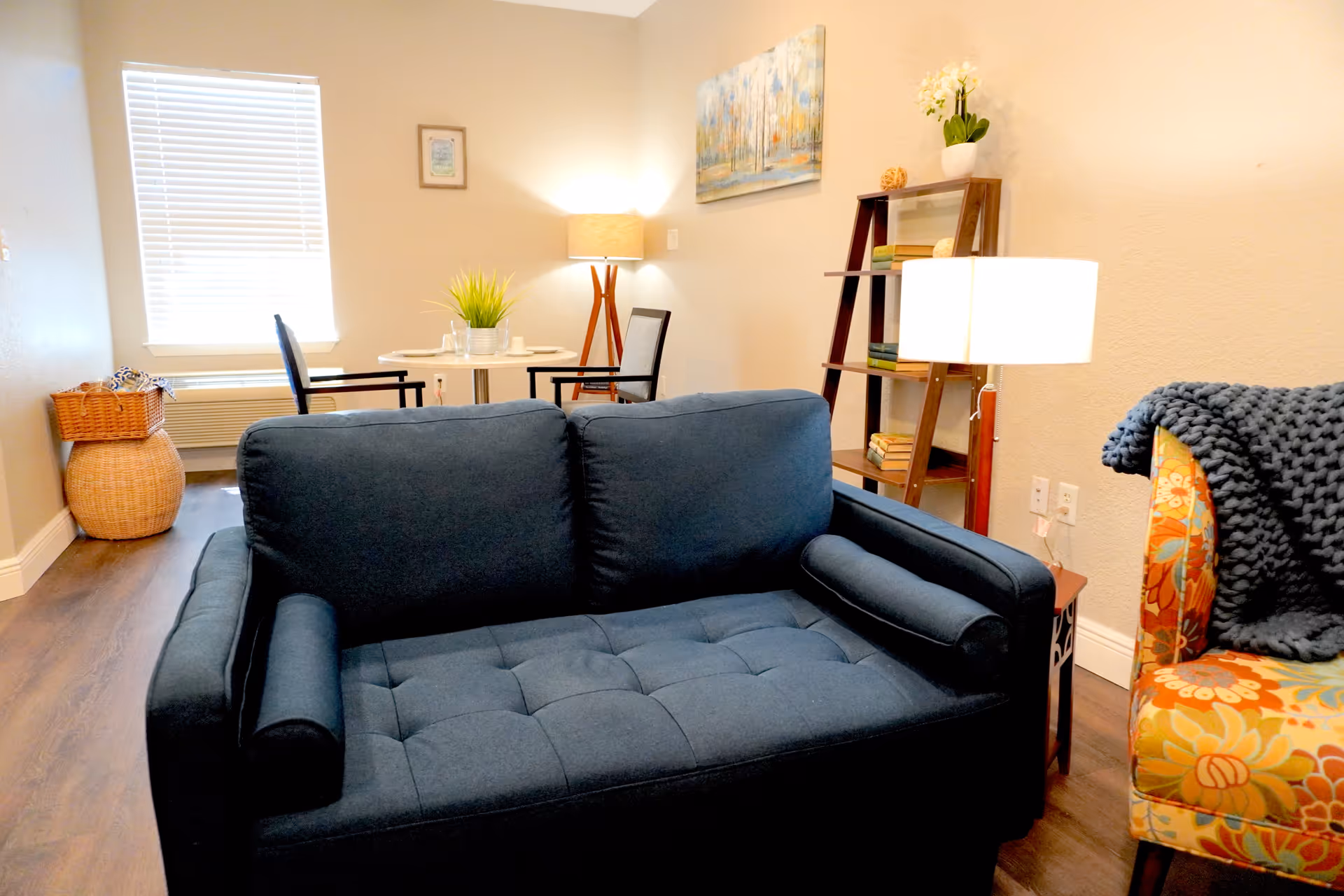 A cozy living room area with a dark blue sofa in the foreground, a colorful floral armchair with a dark knitted throw on the right, a wooden side table with a lamp, a wooden ladder shelf with books and a plant, and a small round dining table with two chairs near a window with blinds.