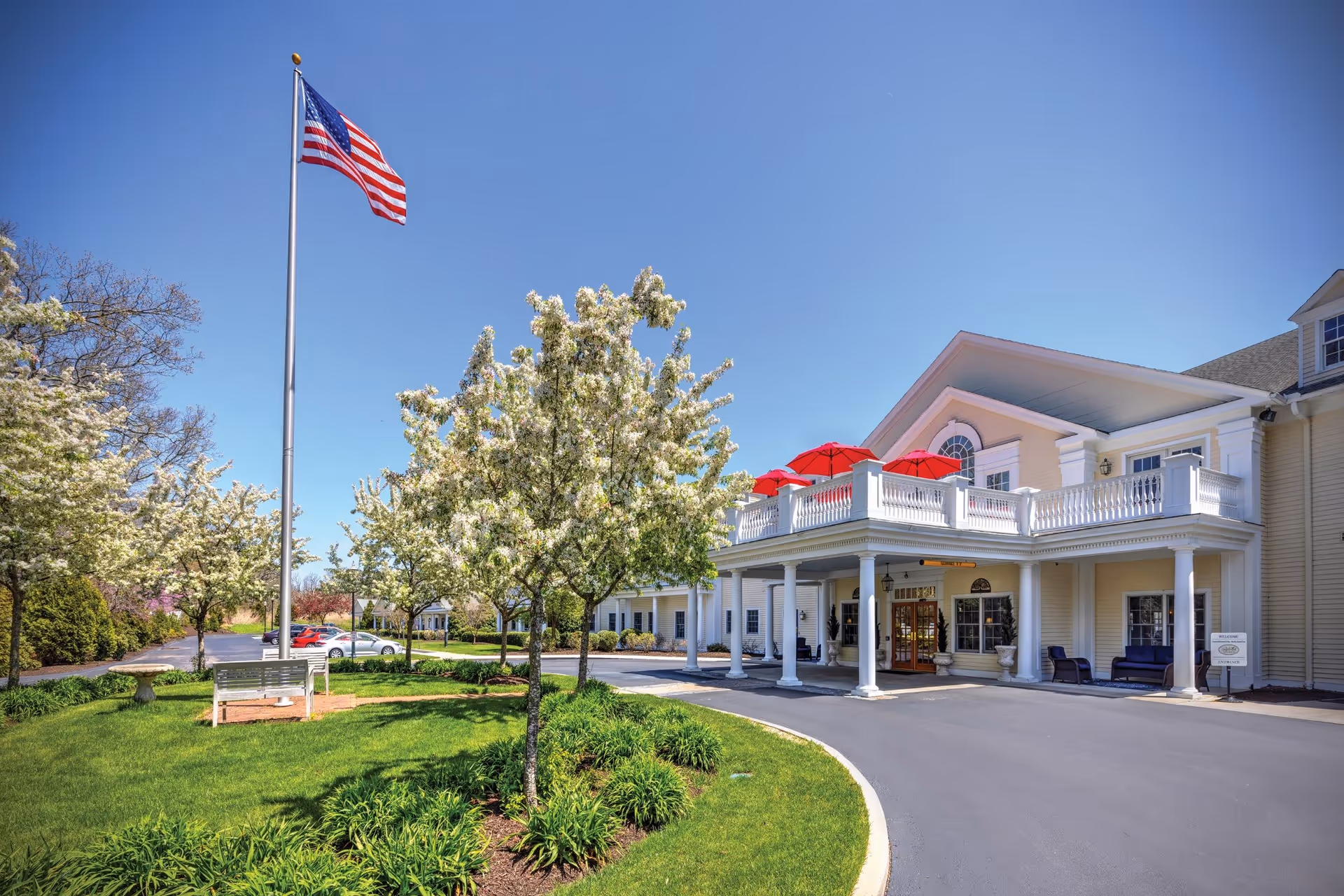Exterior view of The Adel Center building with a covered entrance, white columns, and a balcony with red umbrellas. There are blossoming trees, a green lawn, a flagpole with an American flag, and a parking area with cars under a clear blue sky.