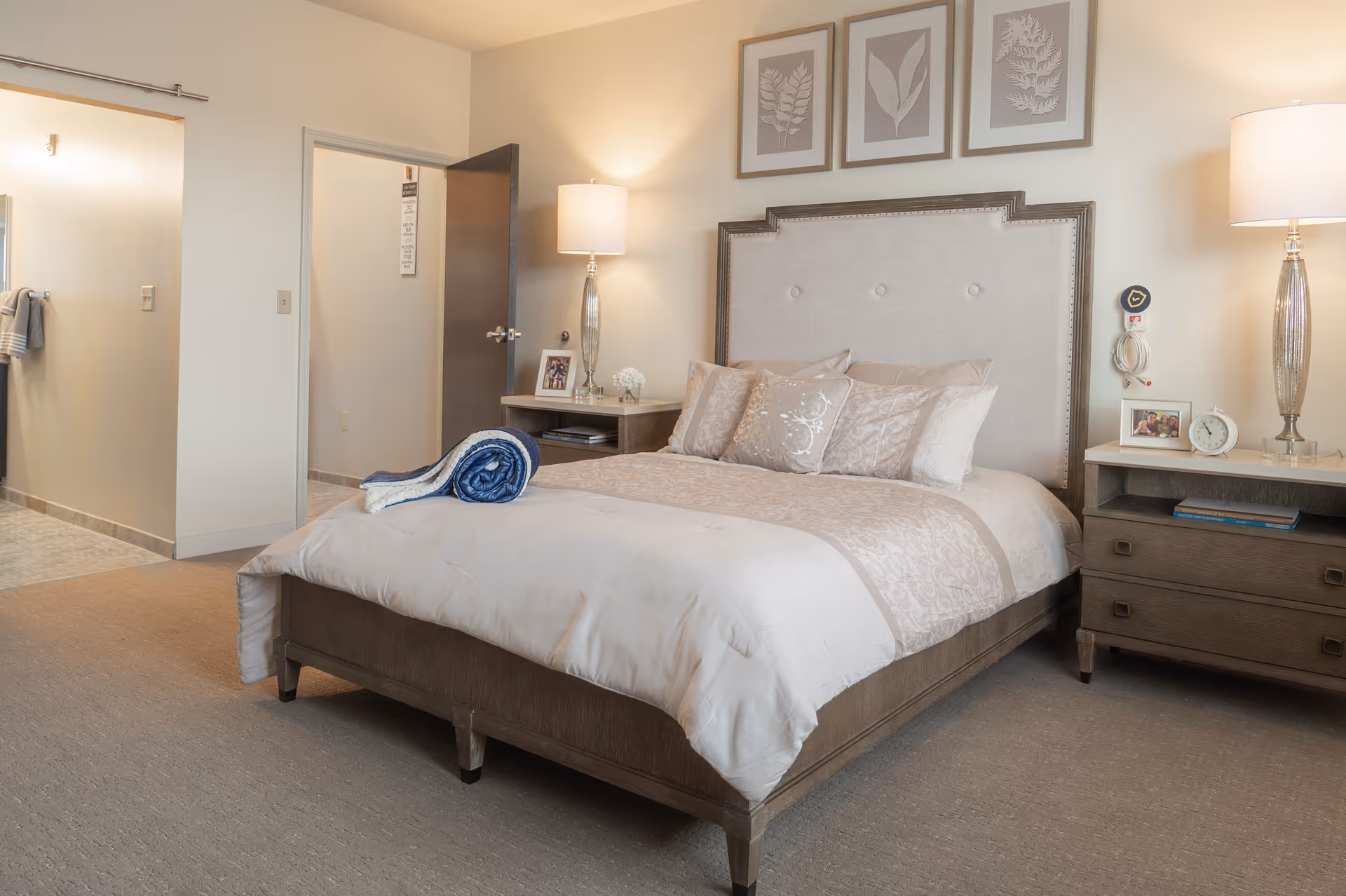 A neatly made bed with a beige and white comforter and multiple pillows in a bedroom. There are two bedside tables with lamps, picture frames, and an alarm clock. The room has beige walls and carpet, with three framed botanical prints above the bed. An open door leads to a hallway and a glimpse of a bathroom with towels hanging on the wall.
