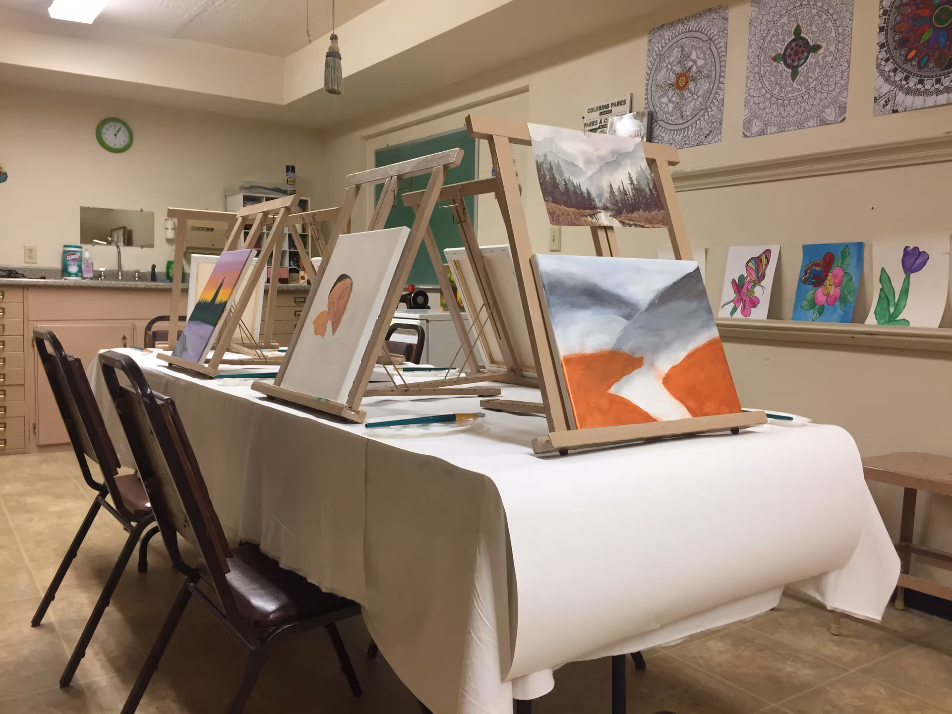 An art room with a table covered in a white cloth, set up with wooden easels holding canvases with various paintings in progress. Chairs are placed around the table. The room has a sink and cabinets in the background, and colorful artwork is displayed on the walls.
