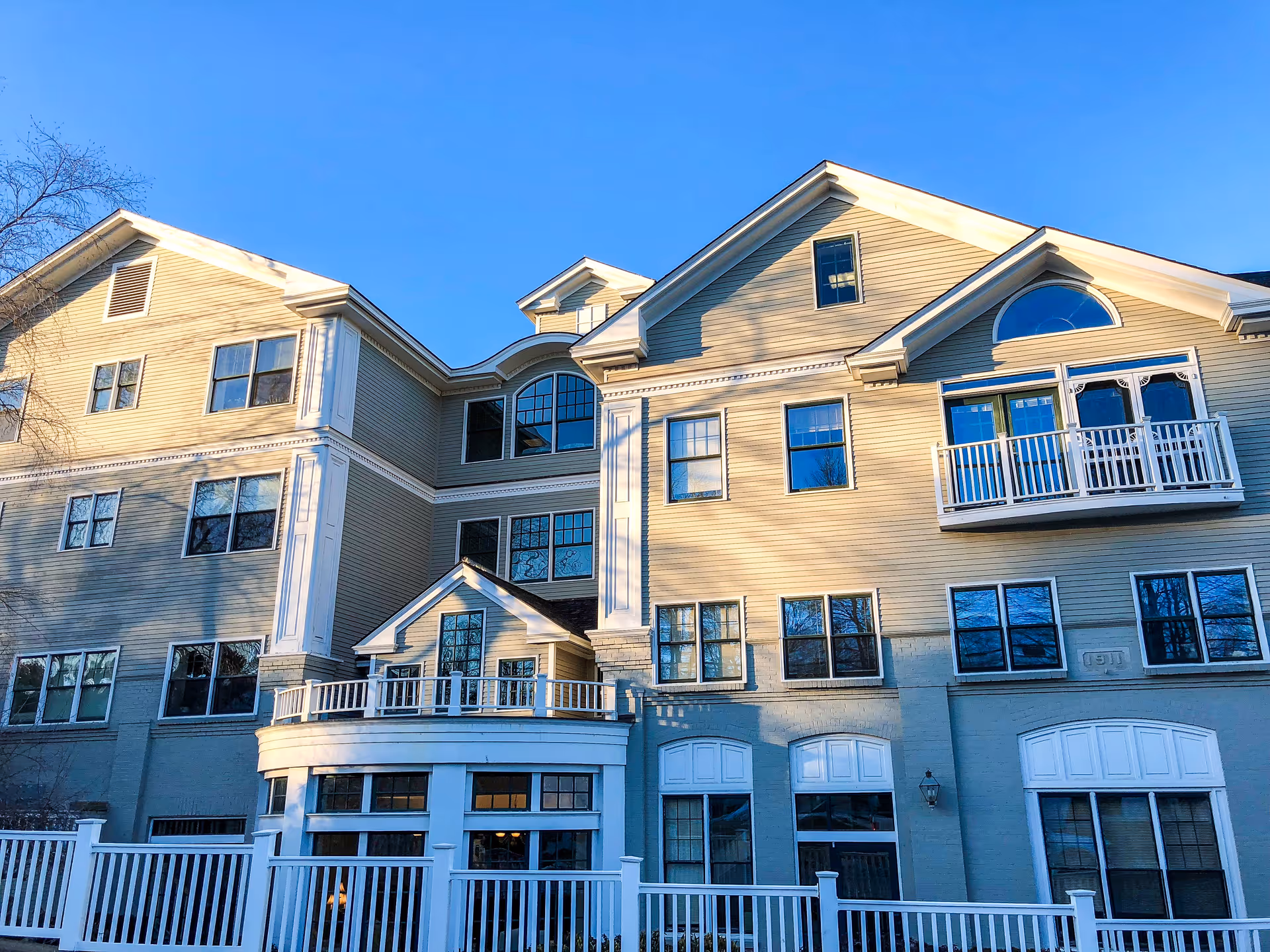 Exterior view of a multi-story senior living facility building with beige siding, multiple windows, a small balcony, and a white fence in front under a clear blue sky.