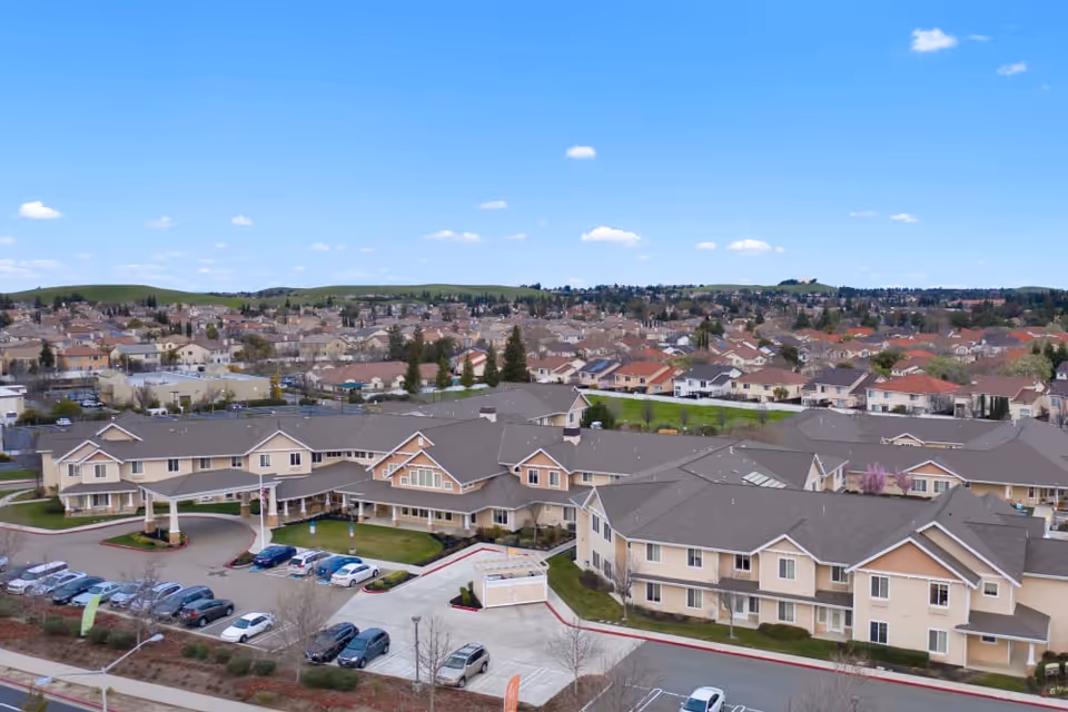 Aerial view of a beige senior living complex with connected buildings, an entrance canopy, parking lot, and surrounding neighborhood.